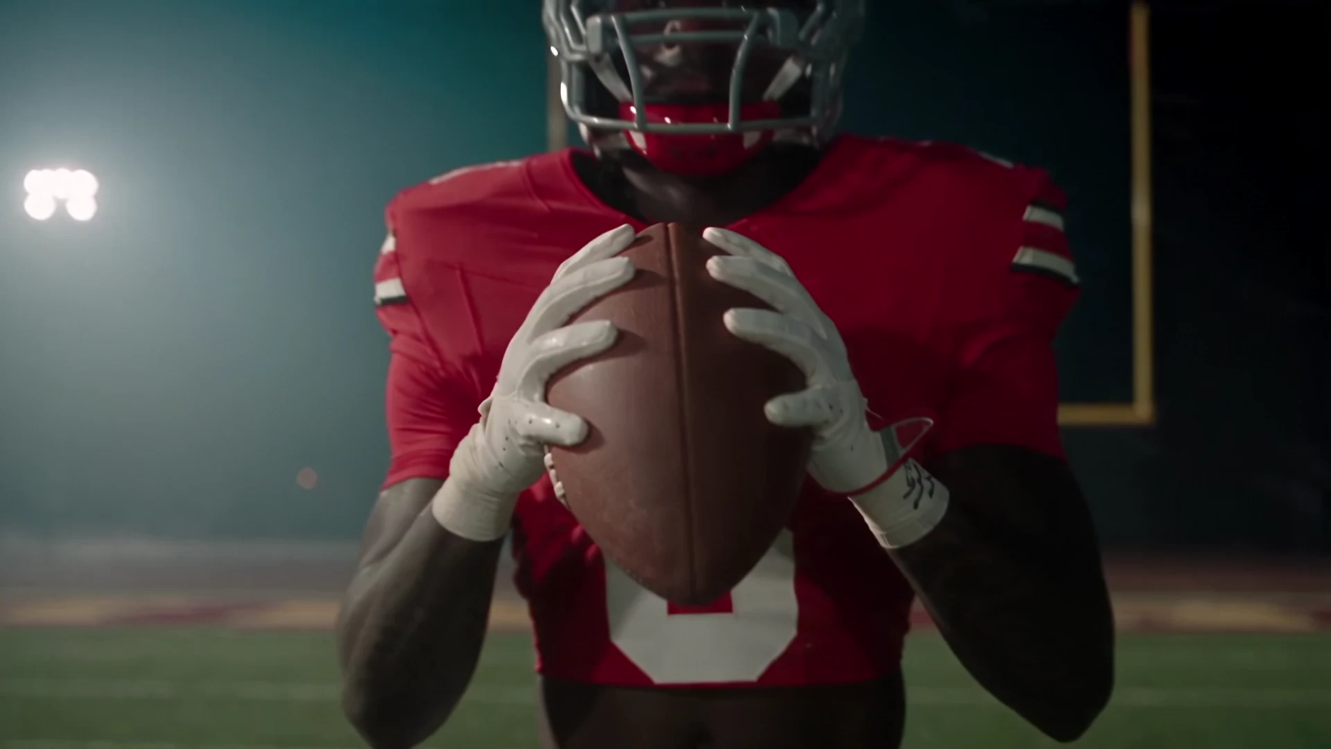 Jaylen McClain in his red No. 8 Ohio State jersey, holding a football at his chest on a dark field at night, stadium lights glowing in the haze and a goal post behind him.